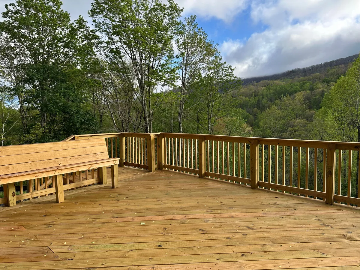 A spacious wooden deck overlooks a stunning mountain vista, surrounded by lush trees. The deck offers comfortable seating, perfect for enjoying the tranquil scenery and fresh mountain air. Early morning sunrise or evening stargazing experiences are suggested by the peaceful setting.