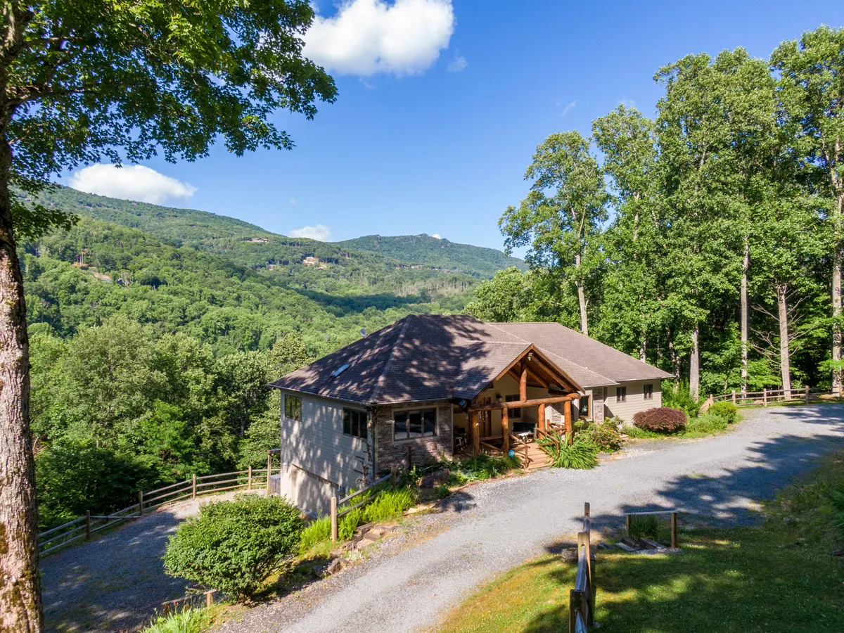 The house is seen from the driveway, framed by lush trees and distant mountains. The wide perspective hints at both the privacy of the setting and the inviting nature of the home, with outdoor spaces for gathering and relaxing.
