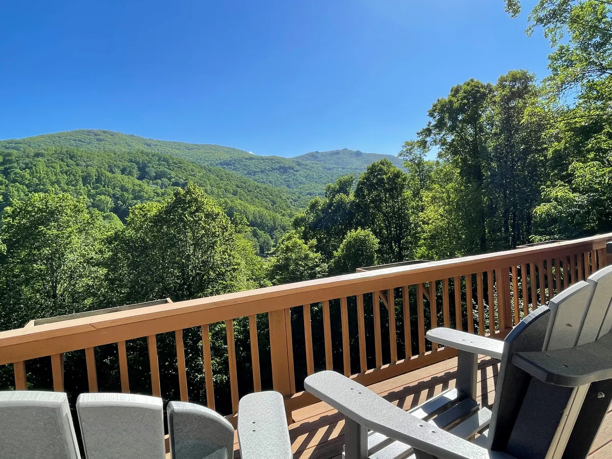 Wooden Adirondack chairs are arranged along a sturdy railing, facing out to a panoramic mountain range. Verdant foliage surrounds the deck, suggesting a peaceful and private setting ideal for contemplation or a casual coffee break.