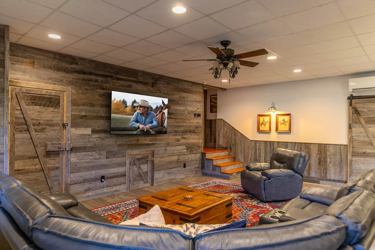 A cozy media lounge with leather couches arranged around a television, a wooden wall, a patterned rug, and a staircase leading to the space. The room exudes a welcoming vibe, perfect for movie marathons or relaxed game nights.