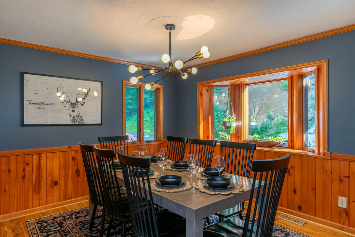 An inviting dining area with a modern table set beneath a stylish light fixture, windows offering glimpses of nature, and elegant artwork, creating a warm setting for shared meals and celebrations.