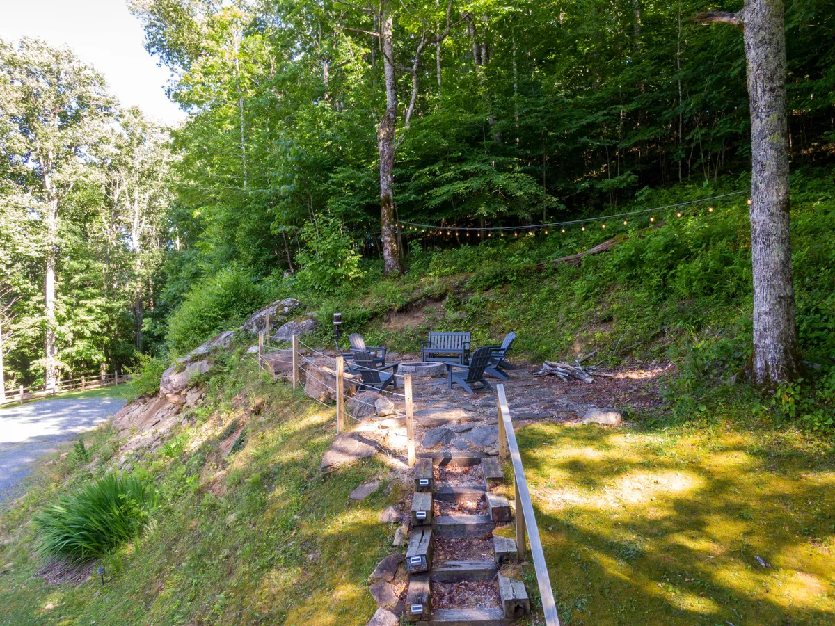A welcoming fire pit encircled by Adirondack chairs sits along a stone pathway, creating a perfect spot for gathering. The natural landscape surrounds the area, offering a sense of seclusion and connection to the outdoors—ideal for storytelling, roasting s’mores, and stargazing under the open sky.
