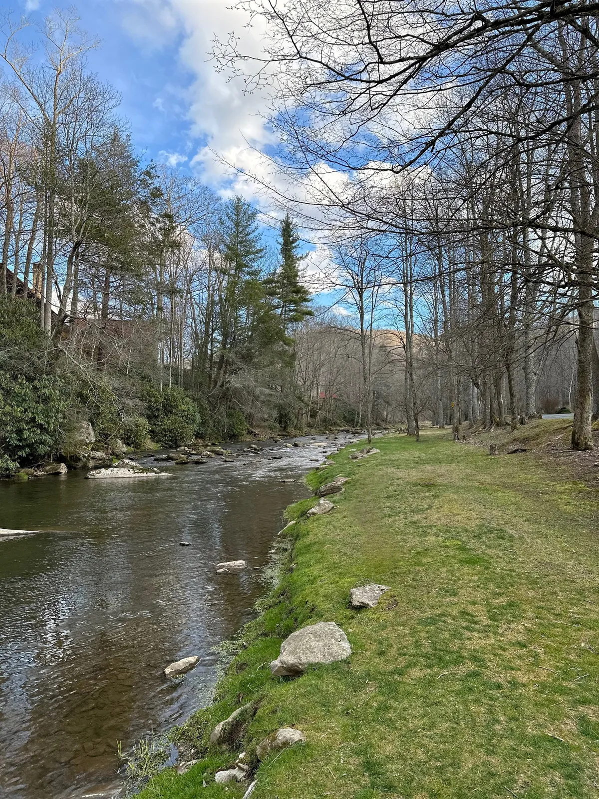 A serene stretch of river winds through trees and grassy banks, with rocks dotting the shoreline. The tranquil water reflects the colors of sunset, suggesting opportunities for fishing, walking, or peaceful reflection.