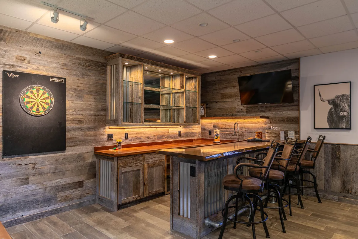 A bar lounge area featuring a dartboard, high stools at the bar counter, glass cabinets with drinkware, and a TV screen. The space is casual and lively, ideal for social gatherings, drinks, and game nights.