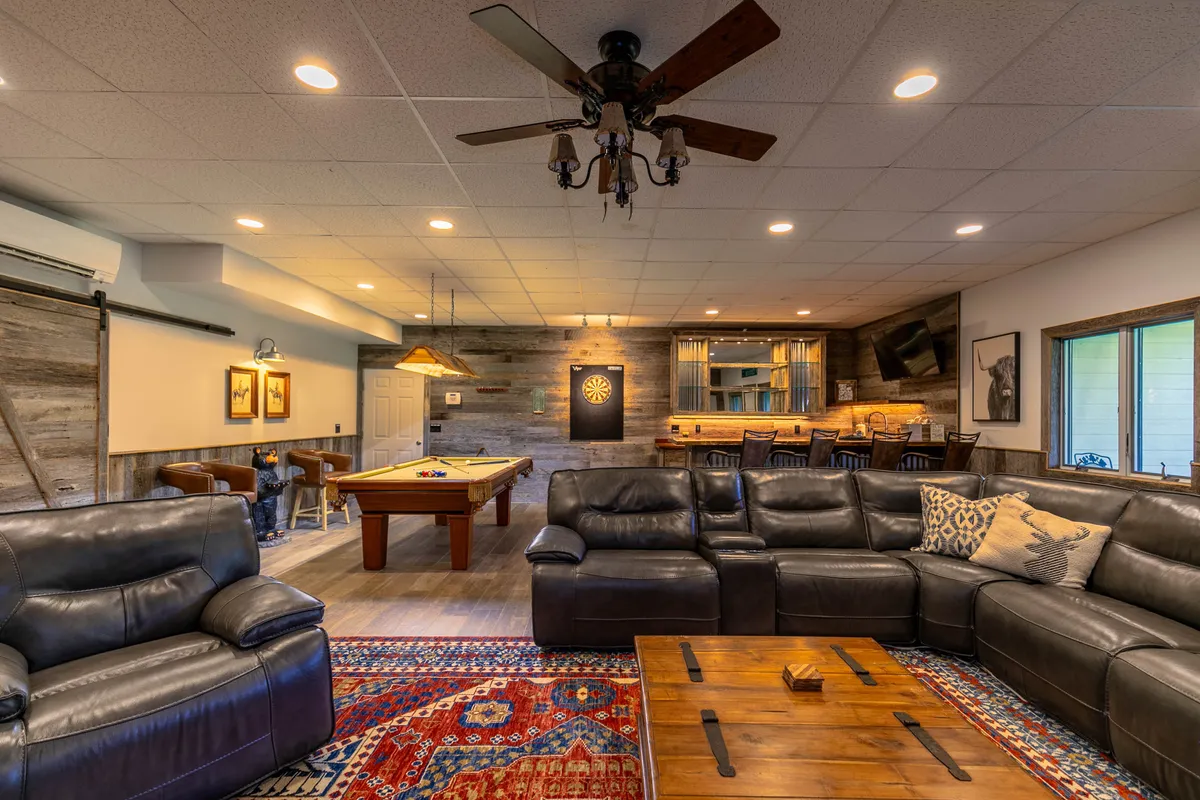 A spacious den featuring leather sofas arranged around a wooden coffee table, a pool table and dartboard in the background, and a wooden bar with stools, all under warm lighting. The room radiates a relaxed, social vibe and clearly serves as a multifunctional entertainment space.