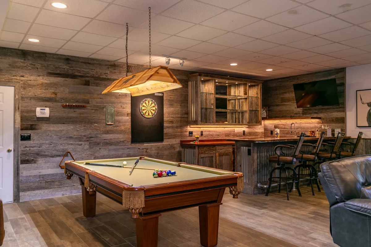 A vibrant entertainment space with a pool table, dartboard, bar area with stools, and a reclaimed wood accent wall. The open layout encourages socializing, while the modern rustic decor adds warmth and character to the room.