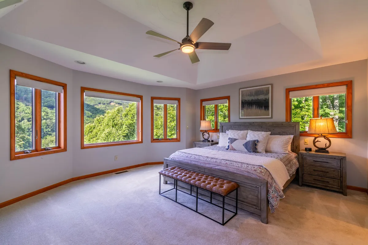 A serene bedroom scene featuring a comfortable bed with decorative pillows, large windows letting in natural light, nightstands, and tasteful decorative items. The windows offer scenic views, enhancing the sense of retreat and relaxation.