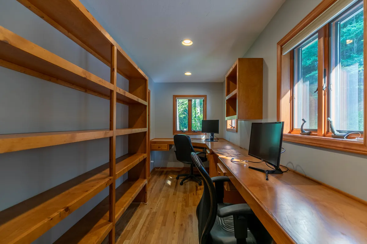 A bright, focused workspace features wooden shelving units, desks equipped with computers, and large windows letting in abundant natural light. The setup is clearly designed for productivity, while the views outside hint at the calm environment beyond.