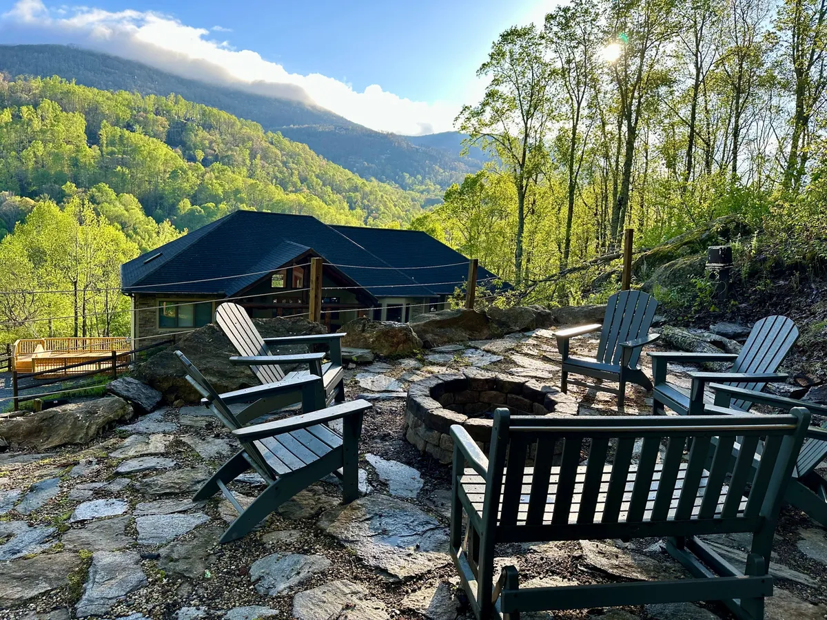 A cozy outdoor fire pit is surrounded by Adirondack chairs, set against vibrant green foliage and mountain views. The scene evokes campfire gatherings, storytelling, and memorable sunset moments.