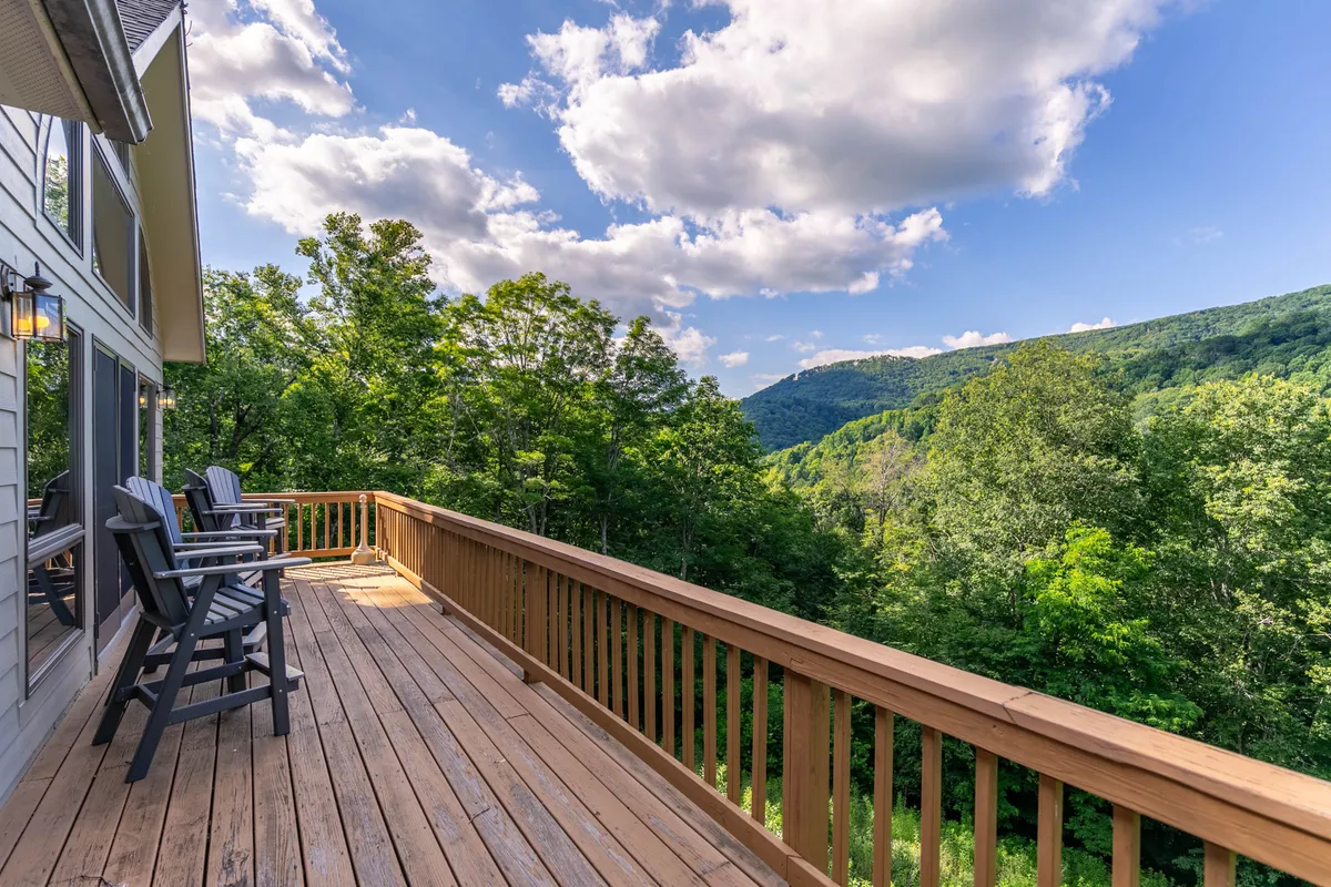 A spacious wooden deck overlooks a stunning mountain view, with lush greenery stretching into the distance under a cloudy sky. The deck is designed for relaxing and soaking in the natural beauty, providing a seamless connection between the home and its serene landscape.