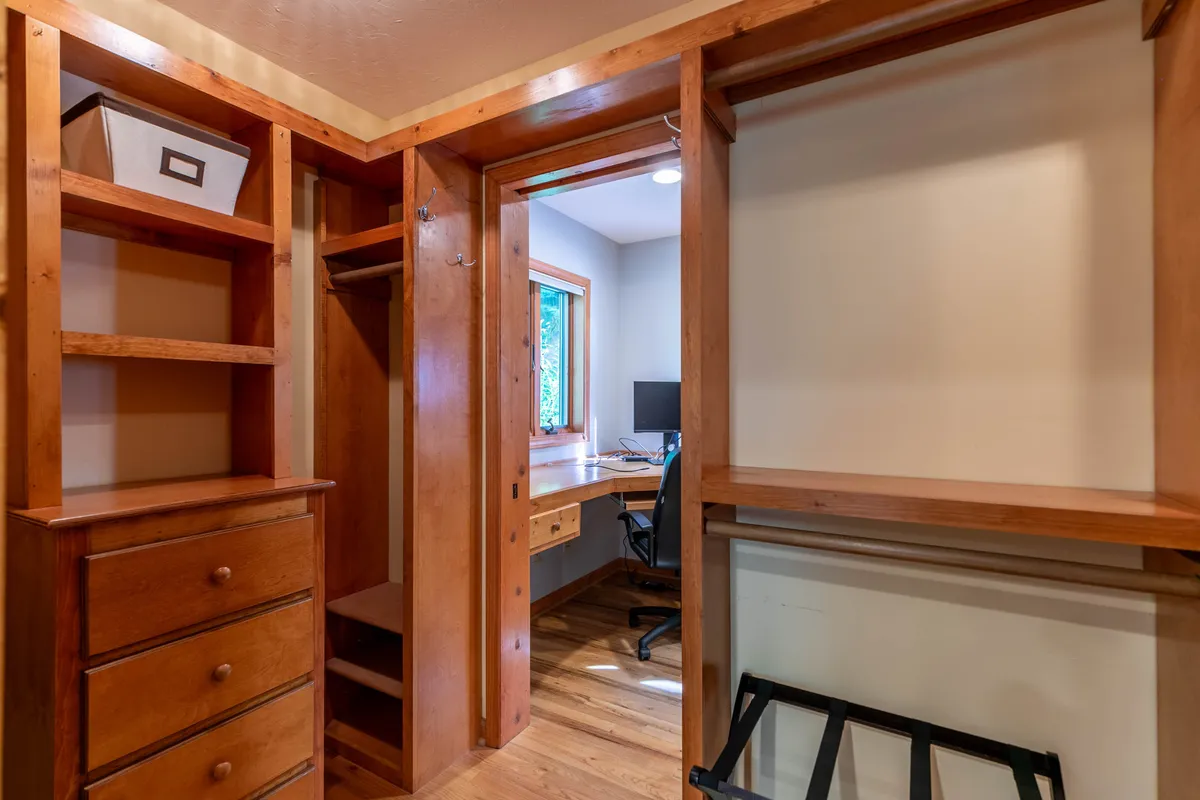 A cozy walk-in closet workspace with wooden shelving, a desktop workspace, and abundant natural light from a window. The atmosphere is productive yet homey, ideal for remote work or quiet reading.