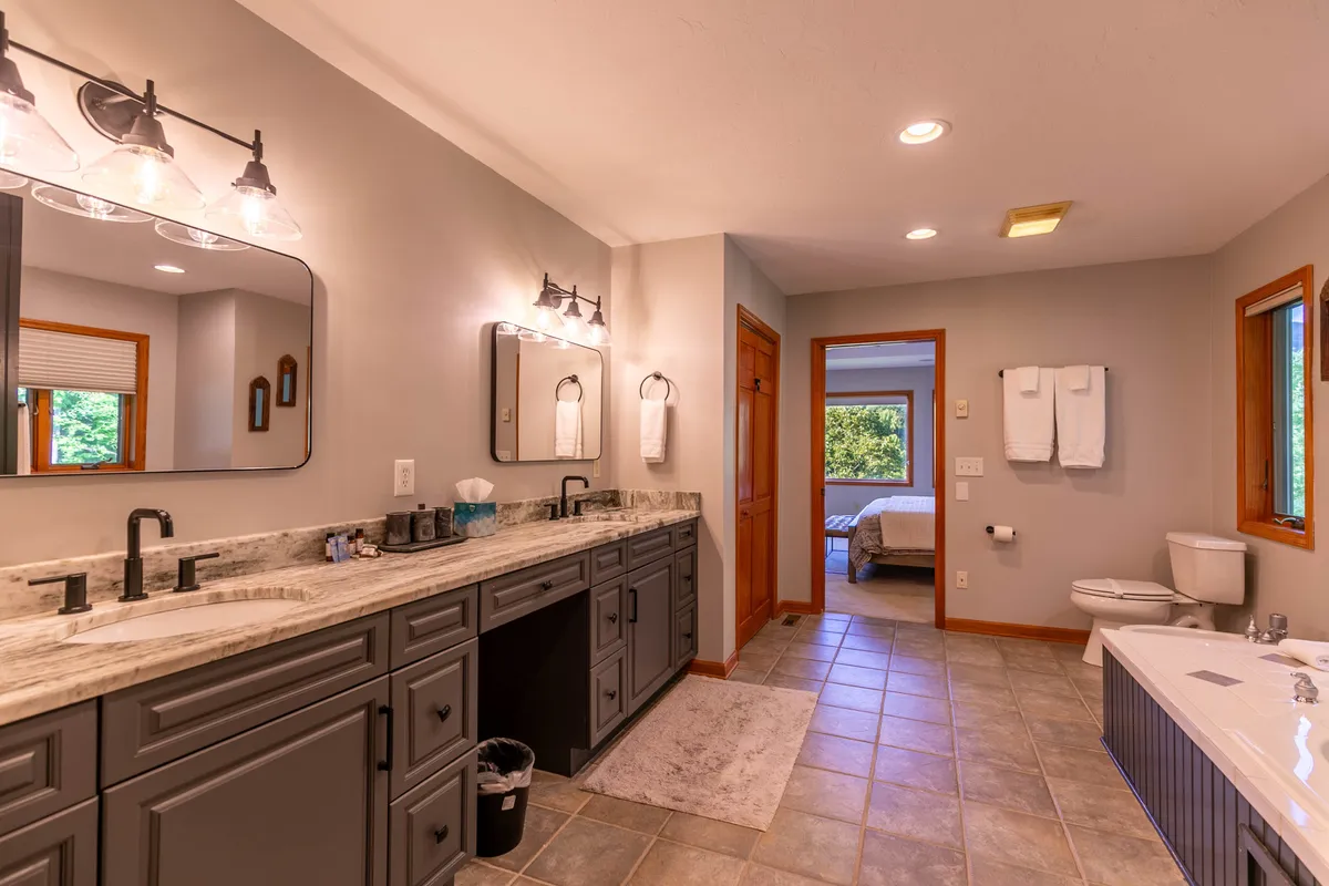 A luxurious bathroom with a double vanity and mirrors, a freestanding bathtub, and a doorway leading to a bedroom. The space is awash with natural light and designed for relaxation and spa-inspired self-care routines.